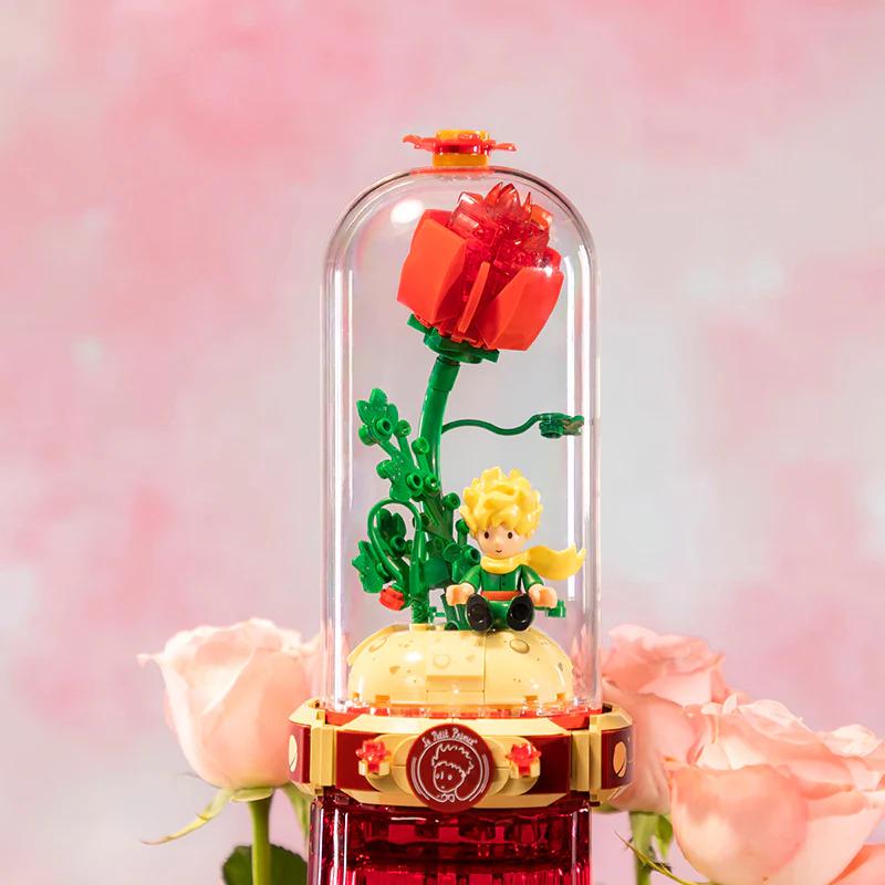 A Lego figurine of a young boy sitting on a yellow base, surrounded by a glass dome containing a red rose and green leaves, with pink roses in the background.
