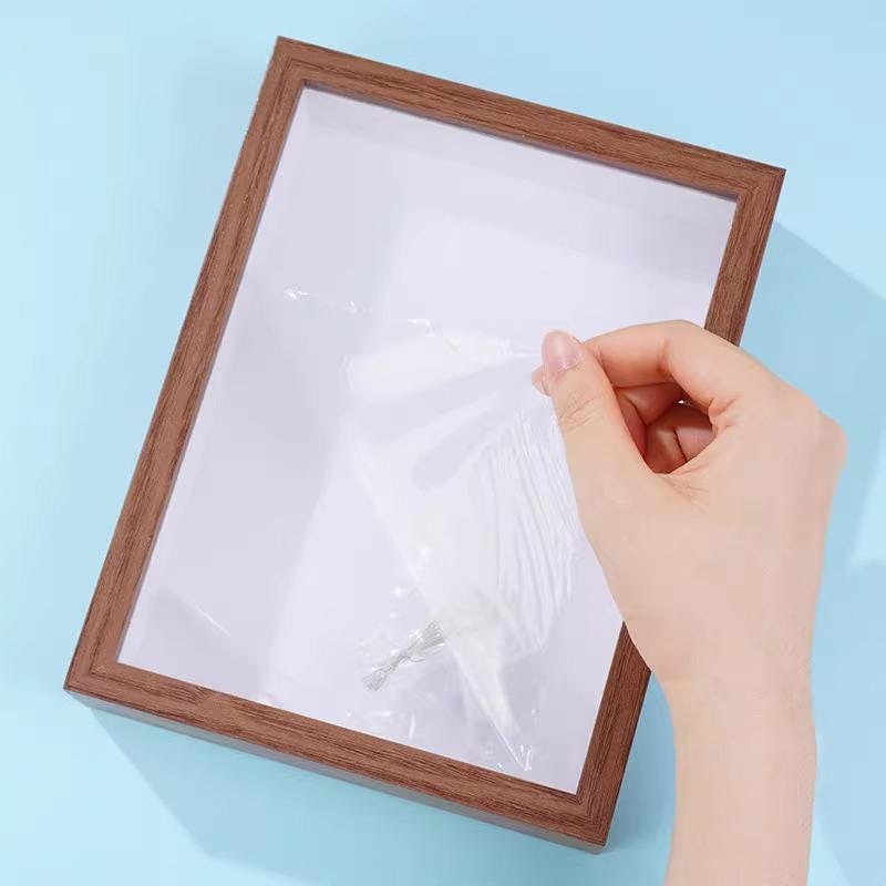 A hand is holding a piece of white paper or a clear plastic sheet, which is partially covering a wooden picture frame.
