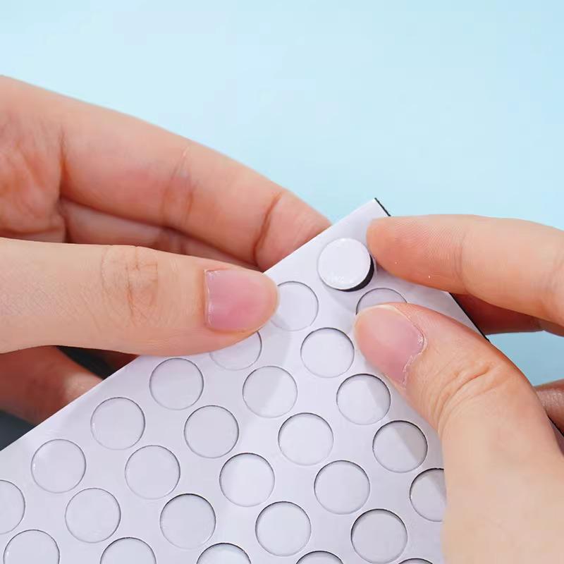 A person's hands are holding a white paper with a grid of circles on it, and they are pressing a small circular object onto the paper.