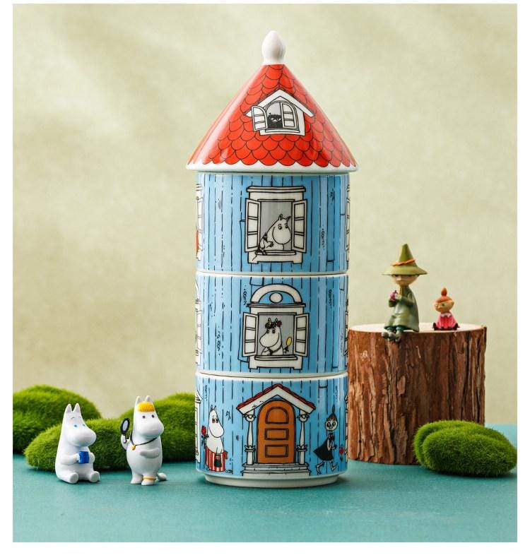 A whimsical ceramic tower with a red roof and blue walls, featuring illustrations of Moomo characters, is surrounded by miniature figurines and a tree stump.