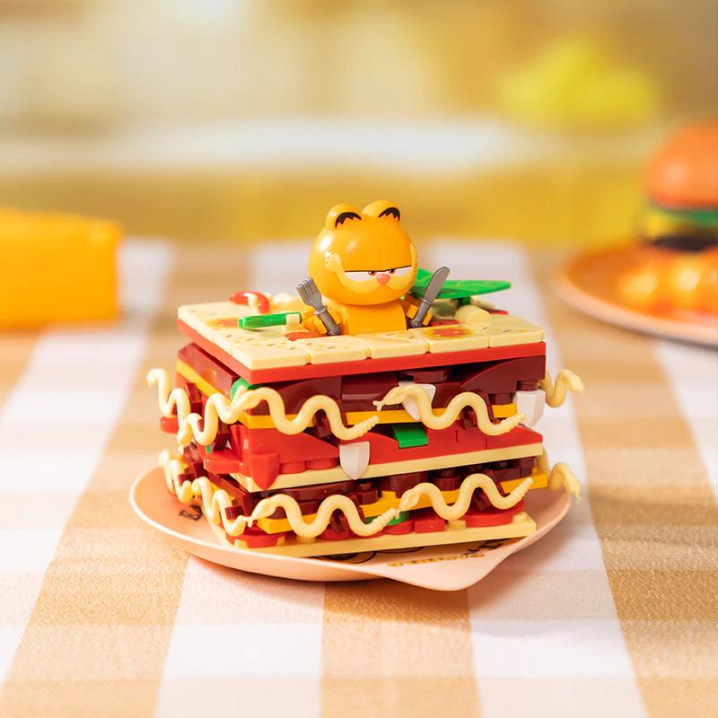 A LEGO figurine of a smiling, cartoon-like character is sitting atop a stack of colorful LEGO bricks, which appear to be part of a sandwich or burger.