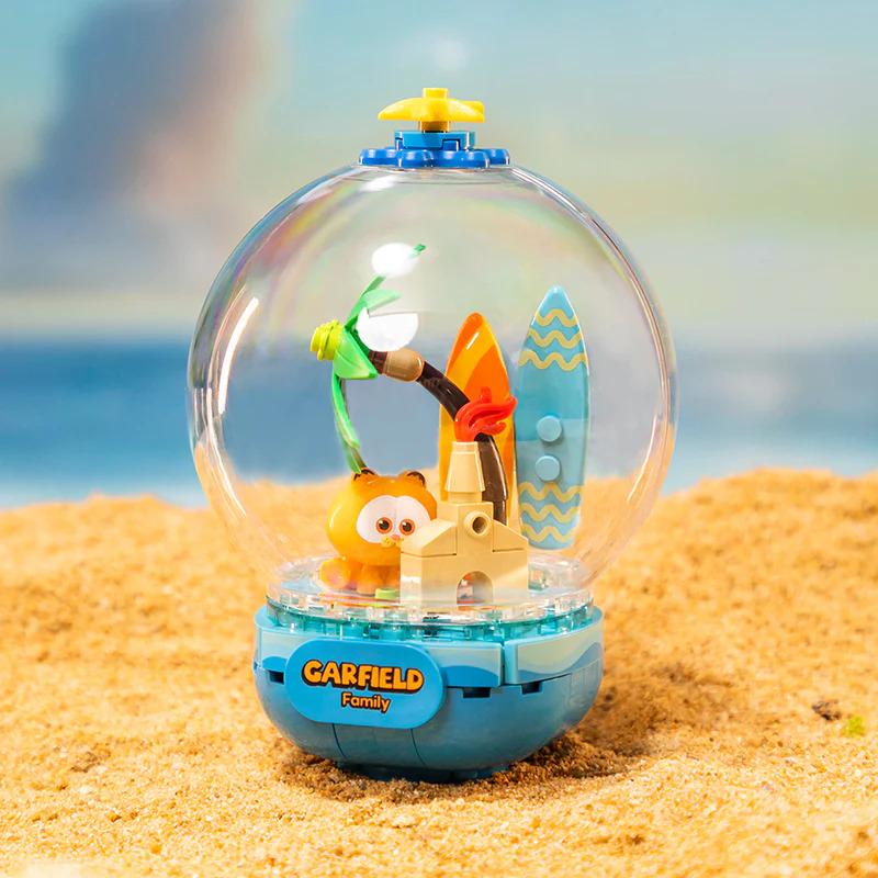 A toy figurine of Garfield, a popular cartoon character, is displayed in a glass globe on a sandy beach.