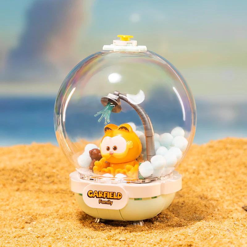 A figurine of a character from the animated series " Garfield" is displayed inside a glass dome, sitting on a sandy surface.