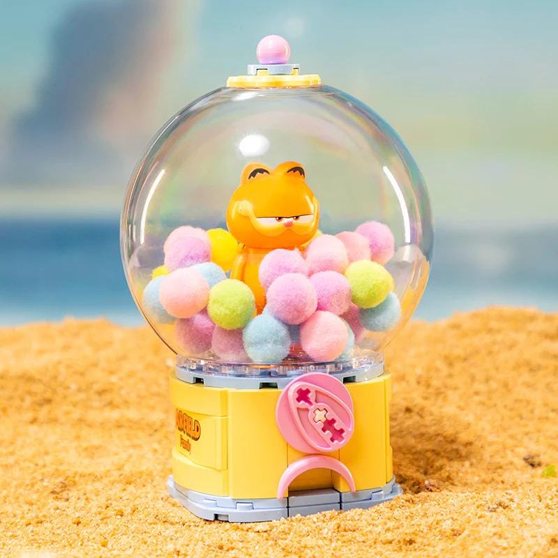 A colorful toy gumball machine filled with multicolored gumballs sits on a sandy beach.