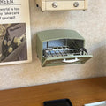 a small green toaster oven on a wooden surface, with a newspaper or magazine visible in the background.