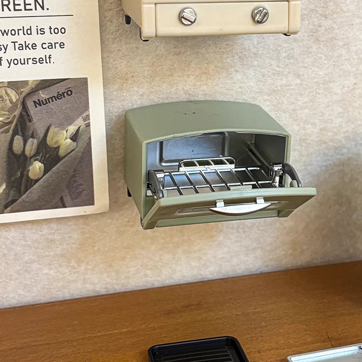 a small green toaster oven on a wooden surface, with a newspaper or magazine visible in the background.