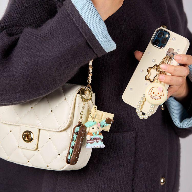 A person is holding a white quilted handbag with a keychain attached to it, and a phone case with a charm attached to it.