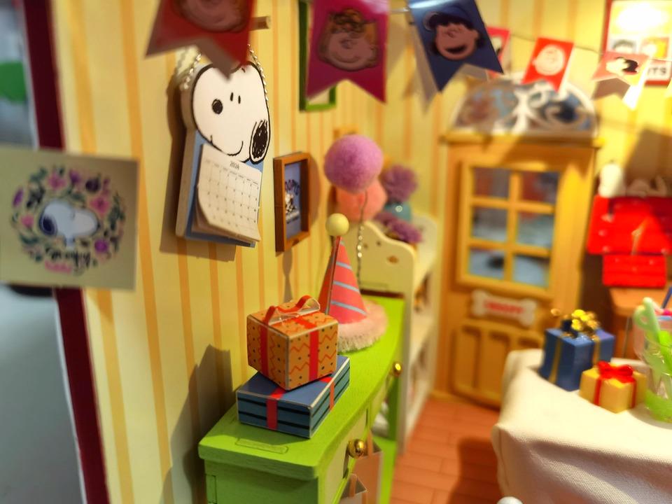 A colorful and whimsical miniature dollhouse filled with various toys and decorations, including a Peanuts-themed wreath, a birthday cake, and a stack of presents.