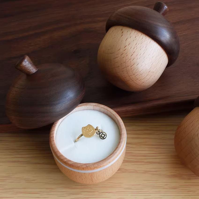 a wooden acorn-shaped container holding a gold ring with a charm, placed on a wooden surface.