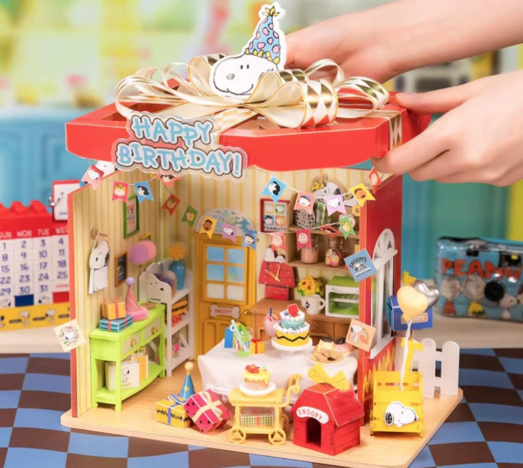 A miniature Peanuts-themed birthday party set, featuring a red box with a "Happy Birthday!" sign, a Snoopy dog toy, and various Peanuts-themed decorations and accessories.