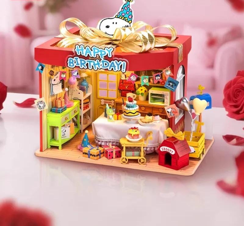 A colorful and whimsical miniature Peanuts-themed dollhouse with a birthday theme, featuring a red roof, a table with a cake, and various Peanuts characters and accessories.