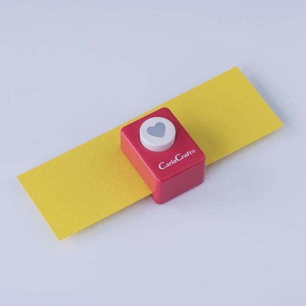 A red and white craft punch with a heart symbol on it, placed on a yellow piece of paper against a white background.