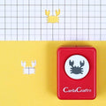 A red hole puncher with a white crab design on it, placed on a yellow background with a grid pattern.