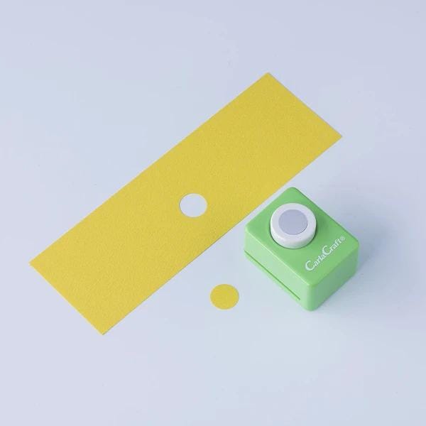 a green push button hole puncher and a yellow piece of paper with a hole in it, both placed on a white background.