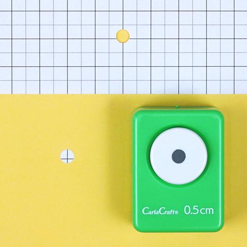 A green push button hole puncher is placed on a yellow background, with a grid pattern visible in the background.