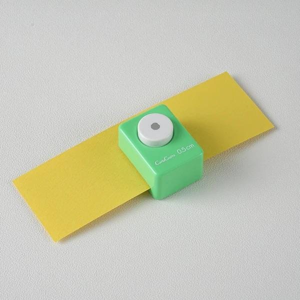 A green push button cutter is placed on a yellow piece of paper against a white background.