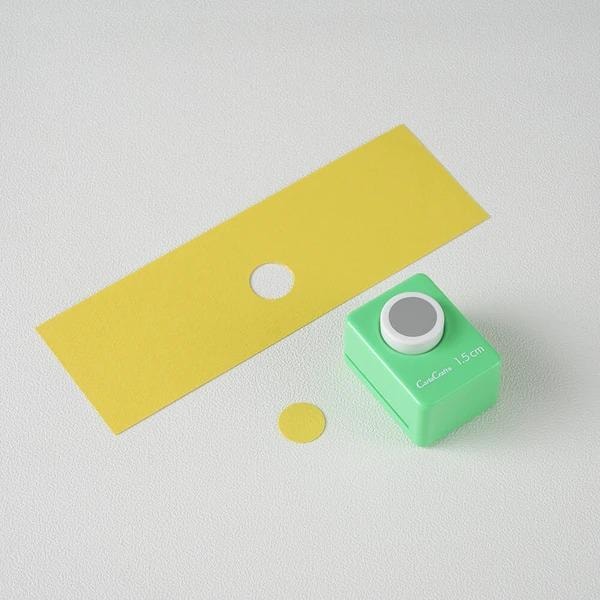 a green push button hole puncher and a yellow piece of paper with a hole in it, both placed on a white surface.
