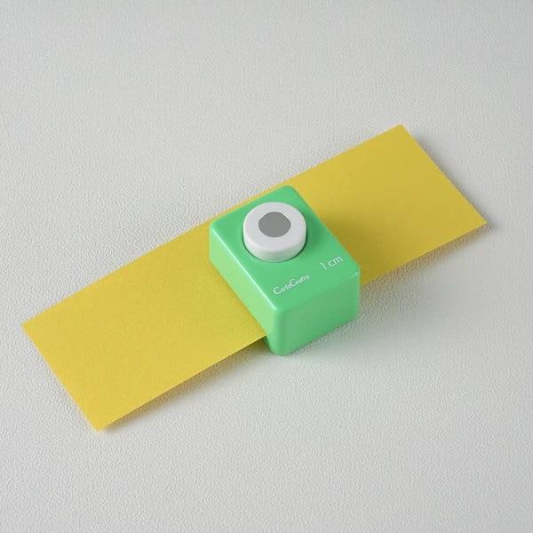 A green push button cutter is placed on a yellow piece of paper against a white background.