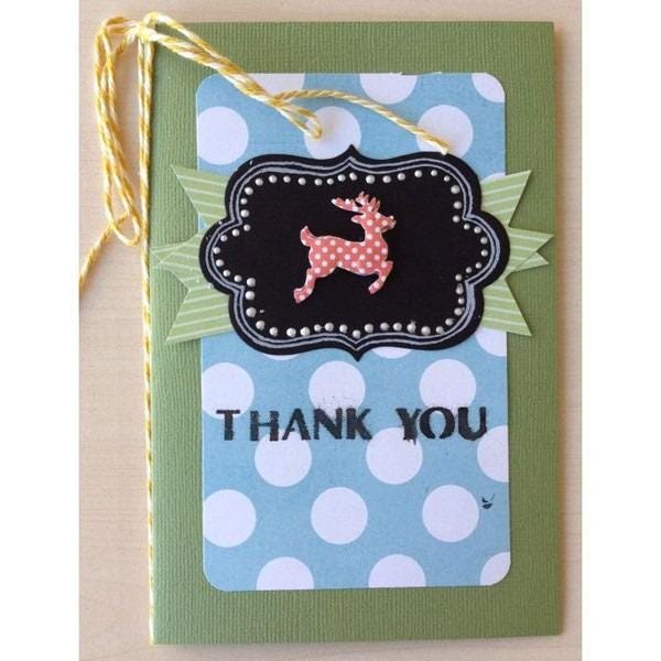 A handmade card with a green background, white polka dots, and a black frame with a red and white polka dot deer. The card has a yellow string tied to it, and the text "THANK YOU" is displayed on the card.