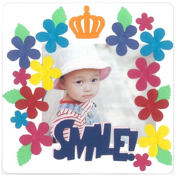 A young child wearing a white hat and striped shirt is surrounded by a colorful frame made of various paper flowers and leaves. The word "SMILE!" is prominently displayed in the center of the frame.