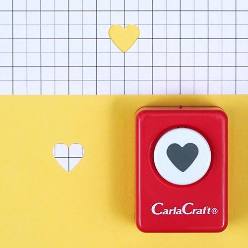 A red hole puncher with a white heart symbol on it, placed on a yellow background with a grid pattern.