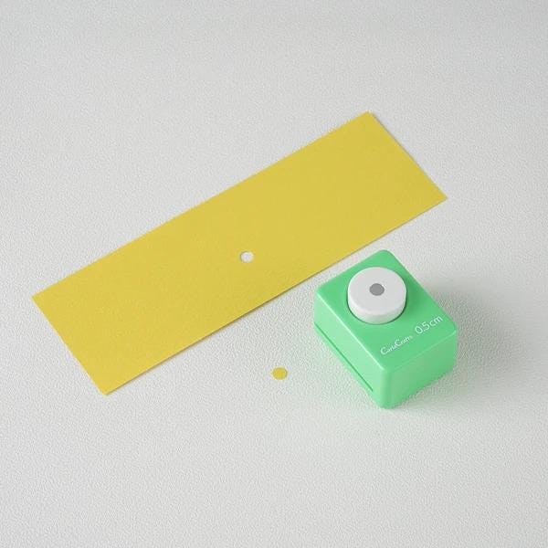 a green push button hole puncher and a yellow piece of paper on a white background.