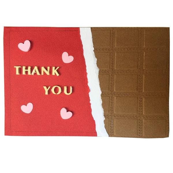 A handmade card with a red background and a chocolate bar on the right side. The card has the text "THANK YOU" written on it, along with three small pink heart shapes scattered around the card.