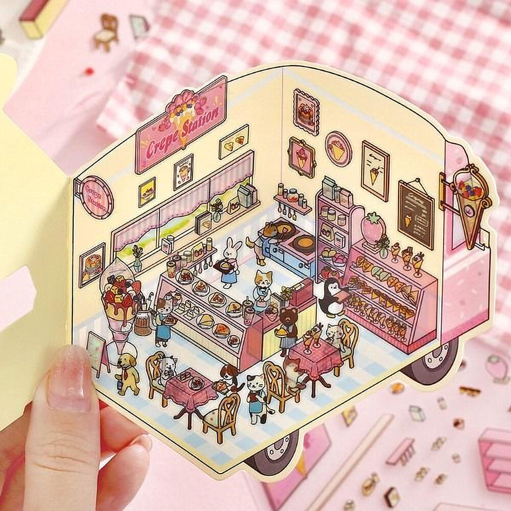 A hand is holding a colorful, illustrated paper cutout depicting a whimsical scene of a crepe shop with various food items and animals.