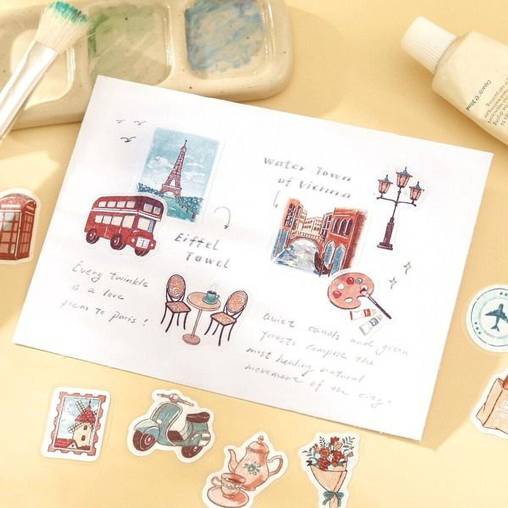 a collection of various objects, including a paper with illustrations of a town, a red double-decker bus, a painting of a bridge, and a teapot, along with some stickers and a brush.