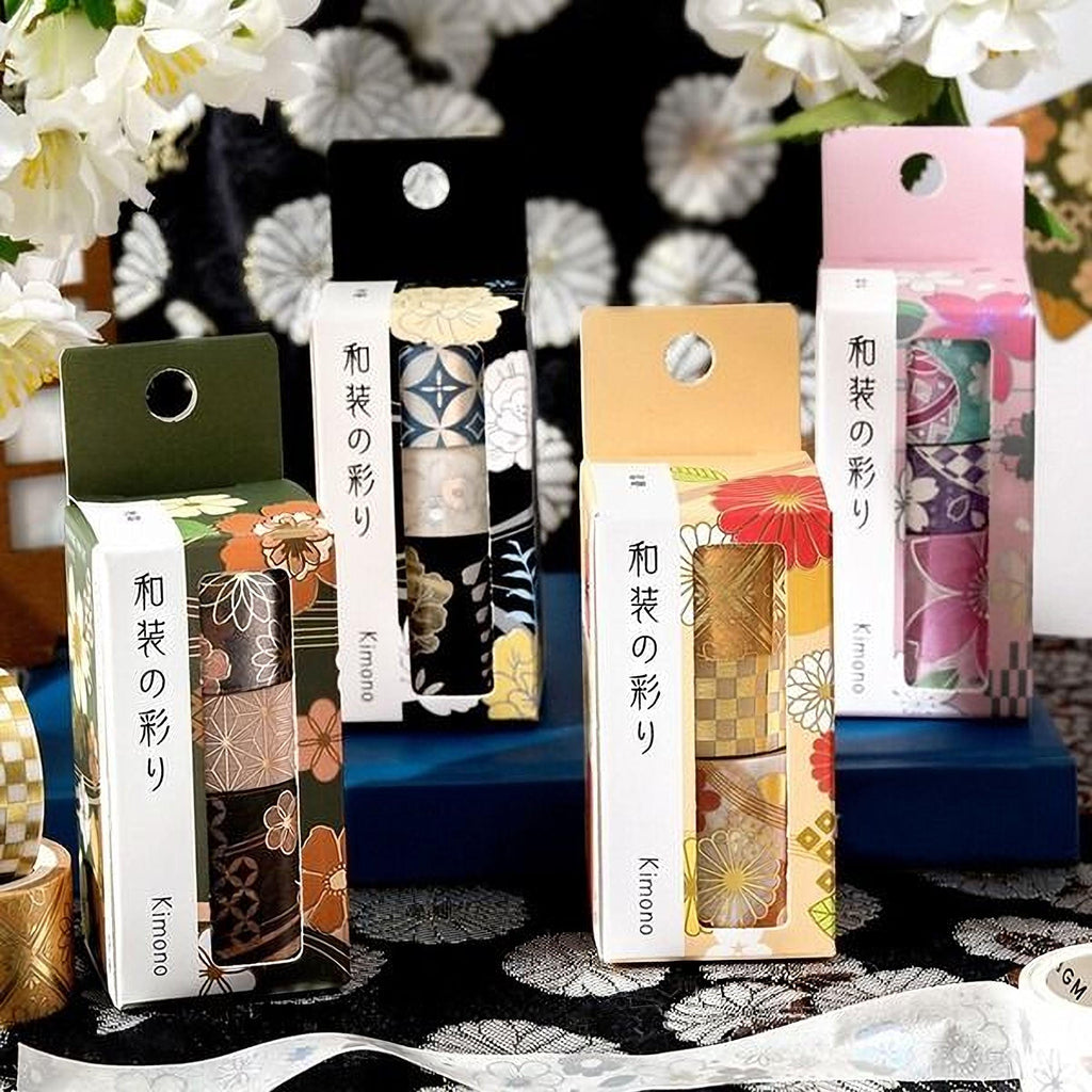 a collection of colorful boxes with Japanese text, likely representing different brands or types of washi tape. The boxes are arranged on a table with a floral patterned tablecloth, and there are white flowers in the background.