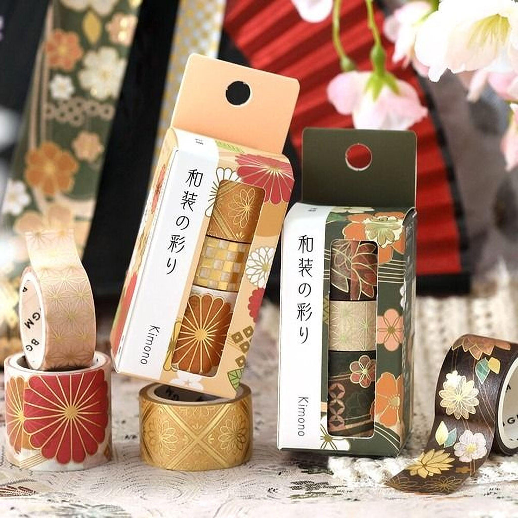 two boxes of decorative tape rolls with Japanese text, placed on a table with additional rolls of tape and flowers in the background.
