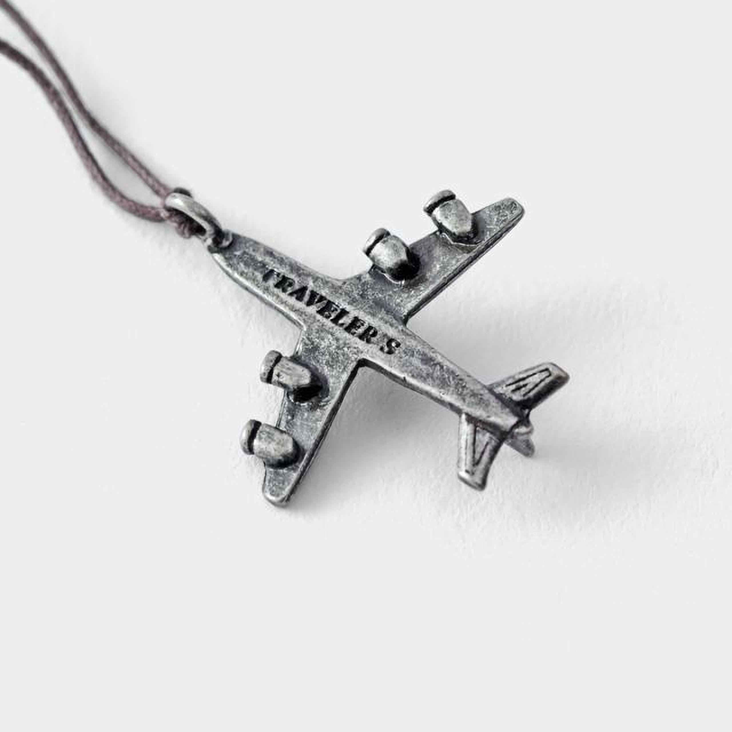 A silver-colored metal airplane pendant with the word "TRAVELERS" engraved on it.