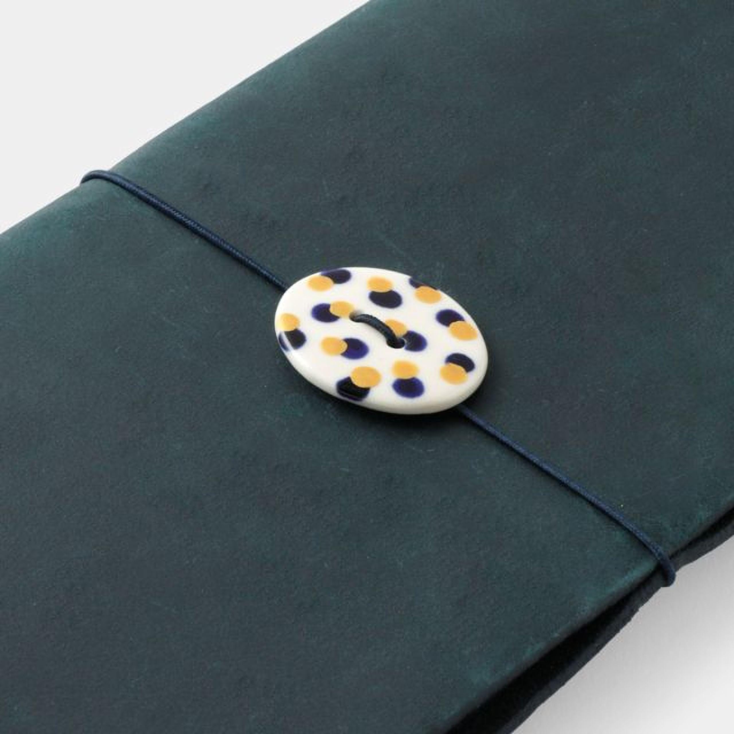 A white button with yellow and blue polka dots is attached to a dark green fabric, which appears to be a piece of leather or a similar material.