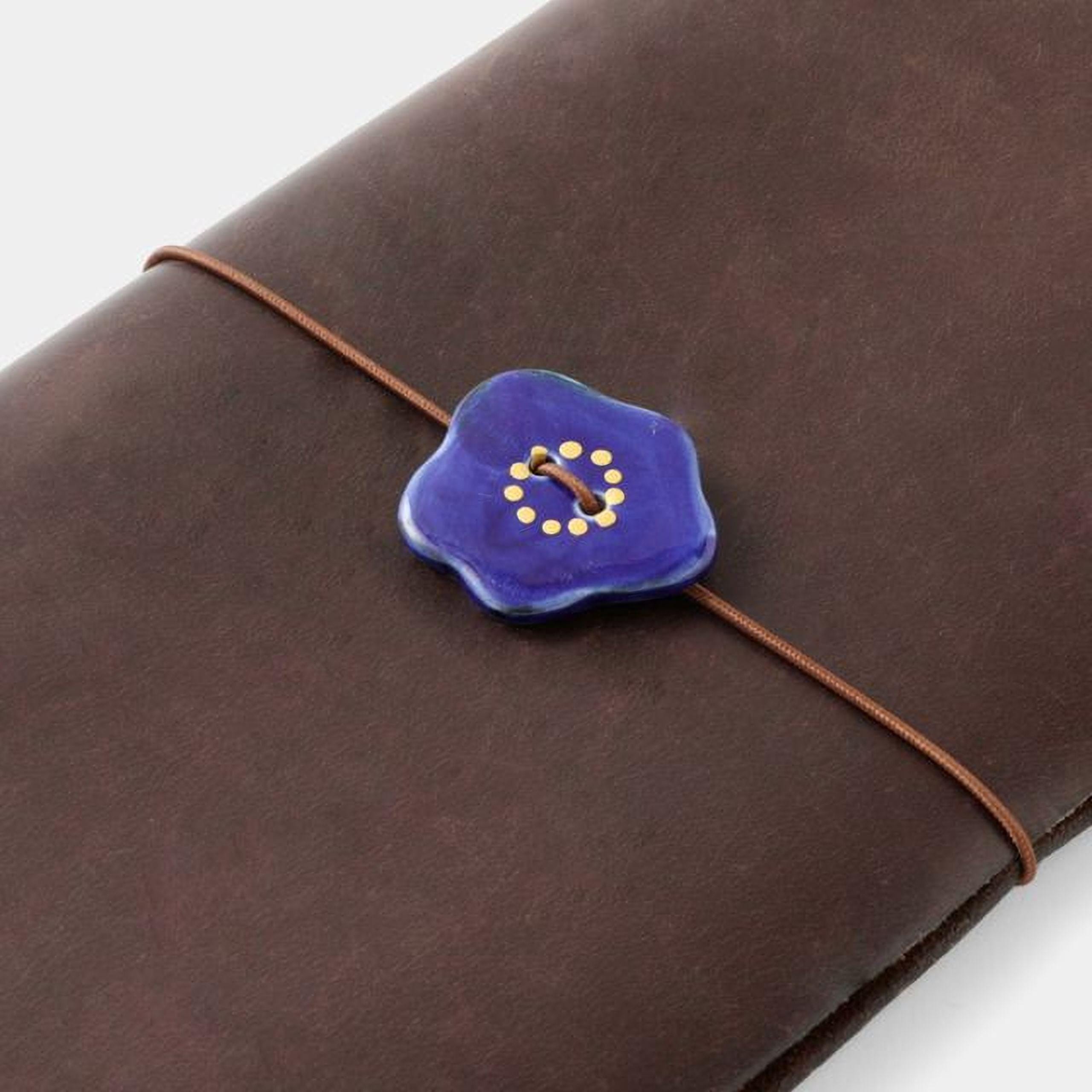 A brown leather-bound notebook with a blue button featuring a yellow floral design on the cover.