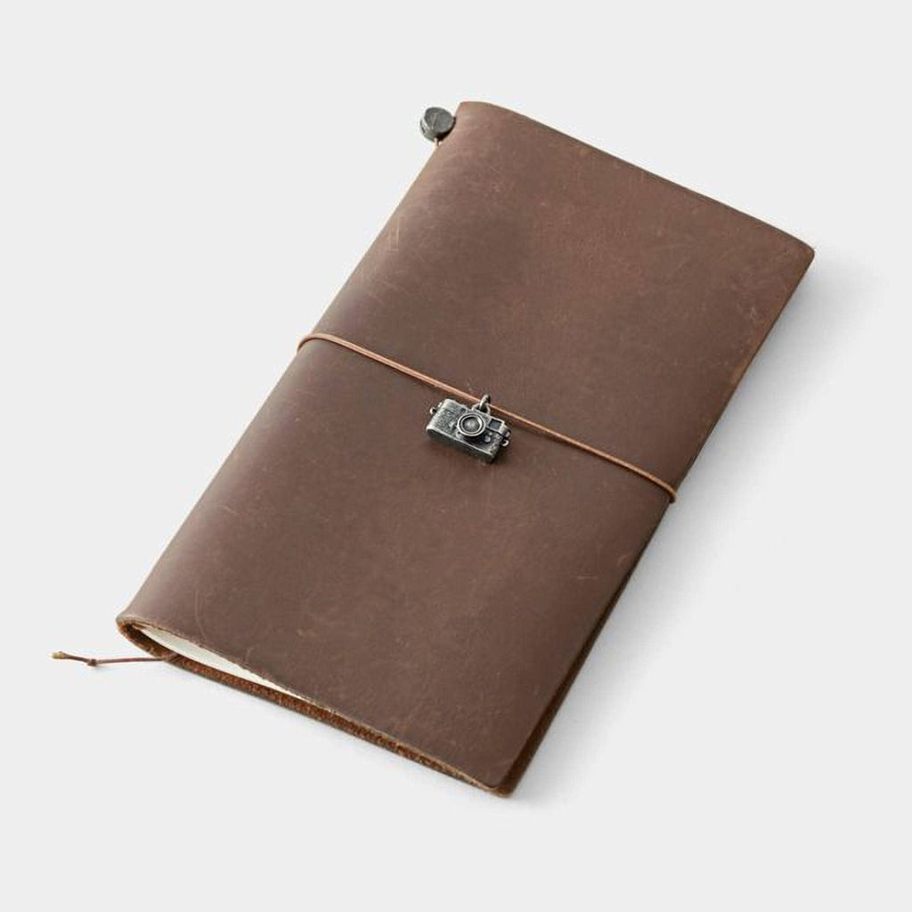 A brown leather-bound notebook with a camera charm attached, resting on a white surface.