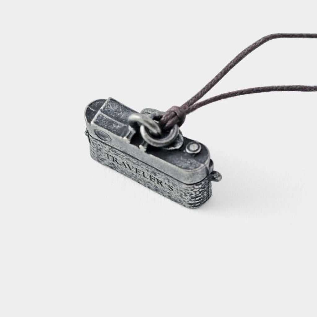 A small metal camera charm with the word "TRAVELERS" engraved on it, attached to a string.