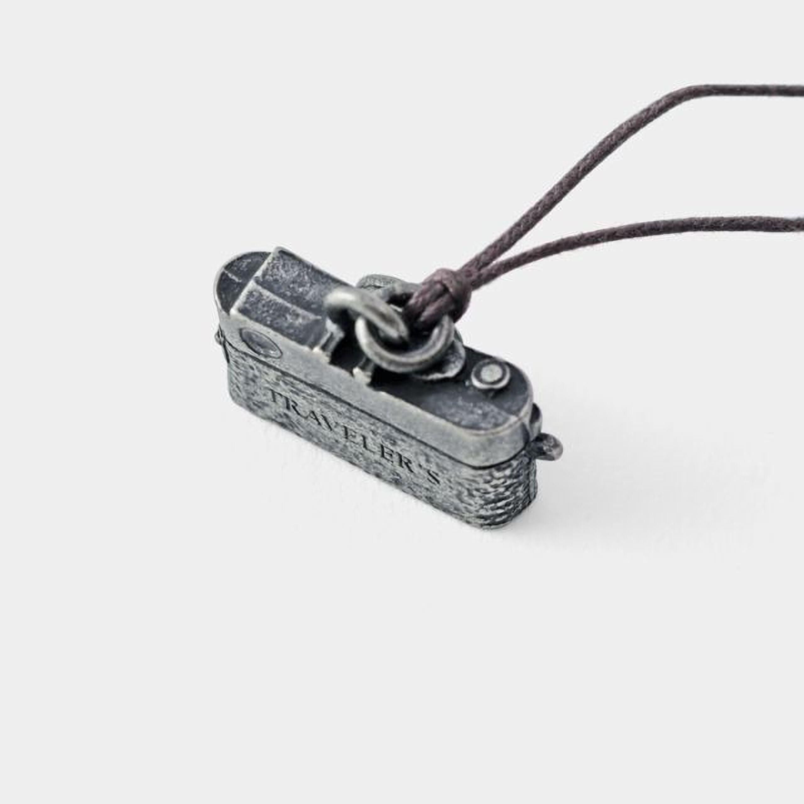 A small metal camera charm with the word "TRAVELERS" engraved on it, attached to a string.