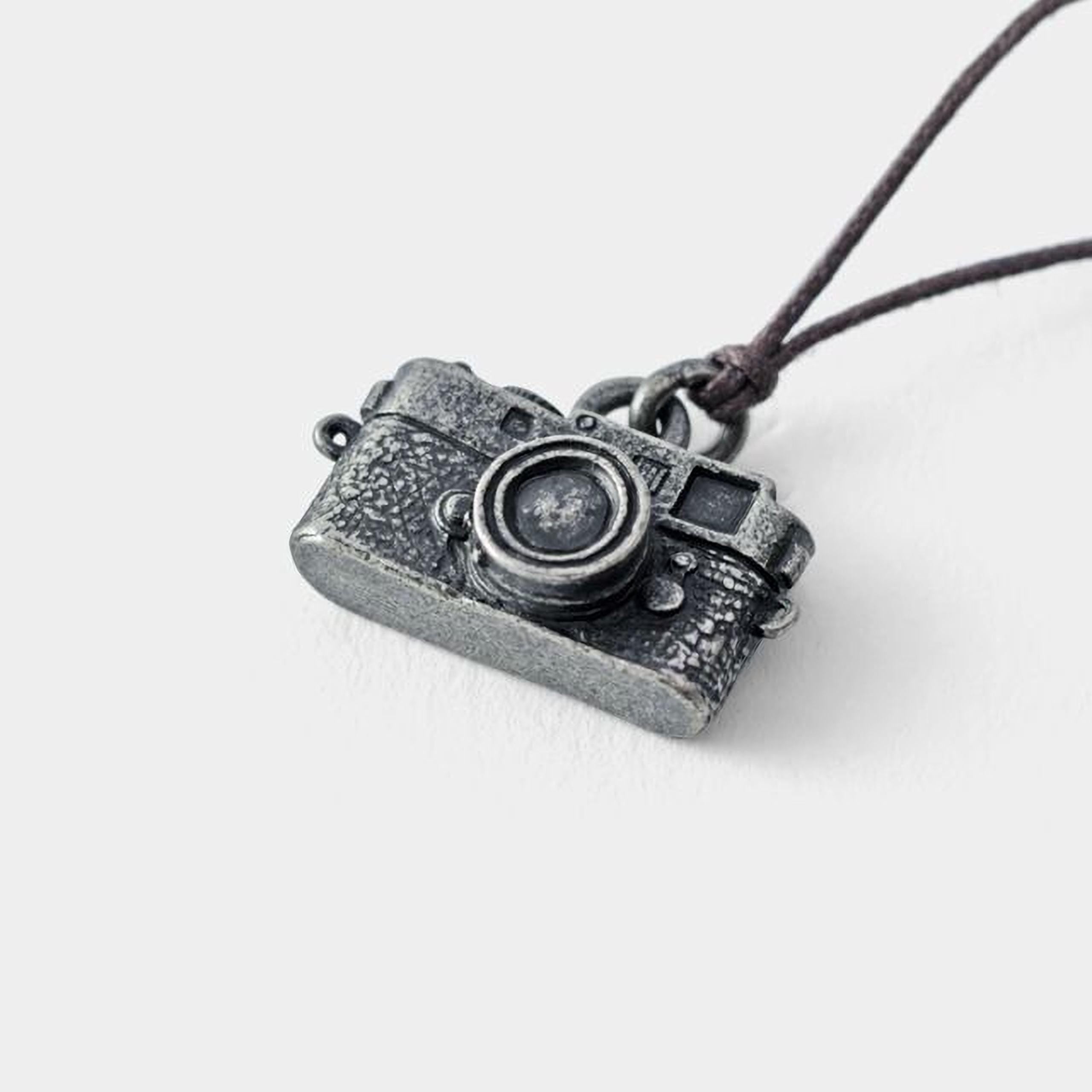 A silver-colored metal pendant in the shape of a camera, hanging from a string.