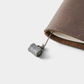 A close-up view of a leather-bound notebook with a metal clip attached to the top edge, resting on a white surface.