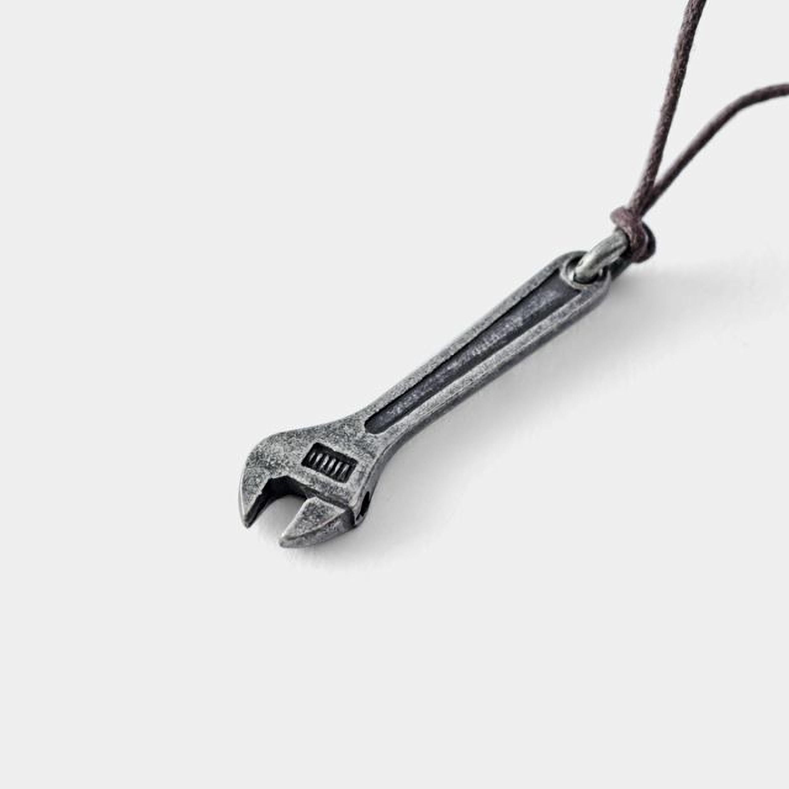 A silver wrench pendant with a leather cord attached, hanging against a plain white background.