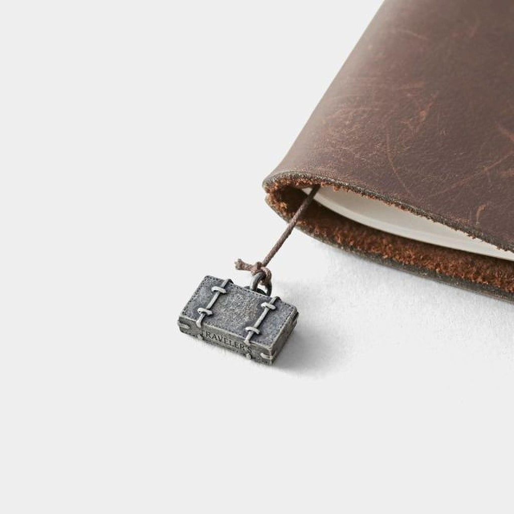 A small metal suitcase charm is attached to a leather-bound notebook.