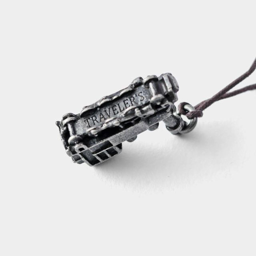 A silver-colored metal charm or pendant with the word "TRAVELER'S" engraved on it, attached to a string.