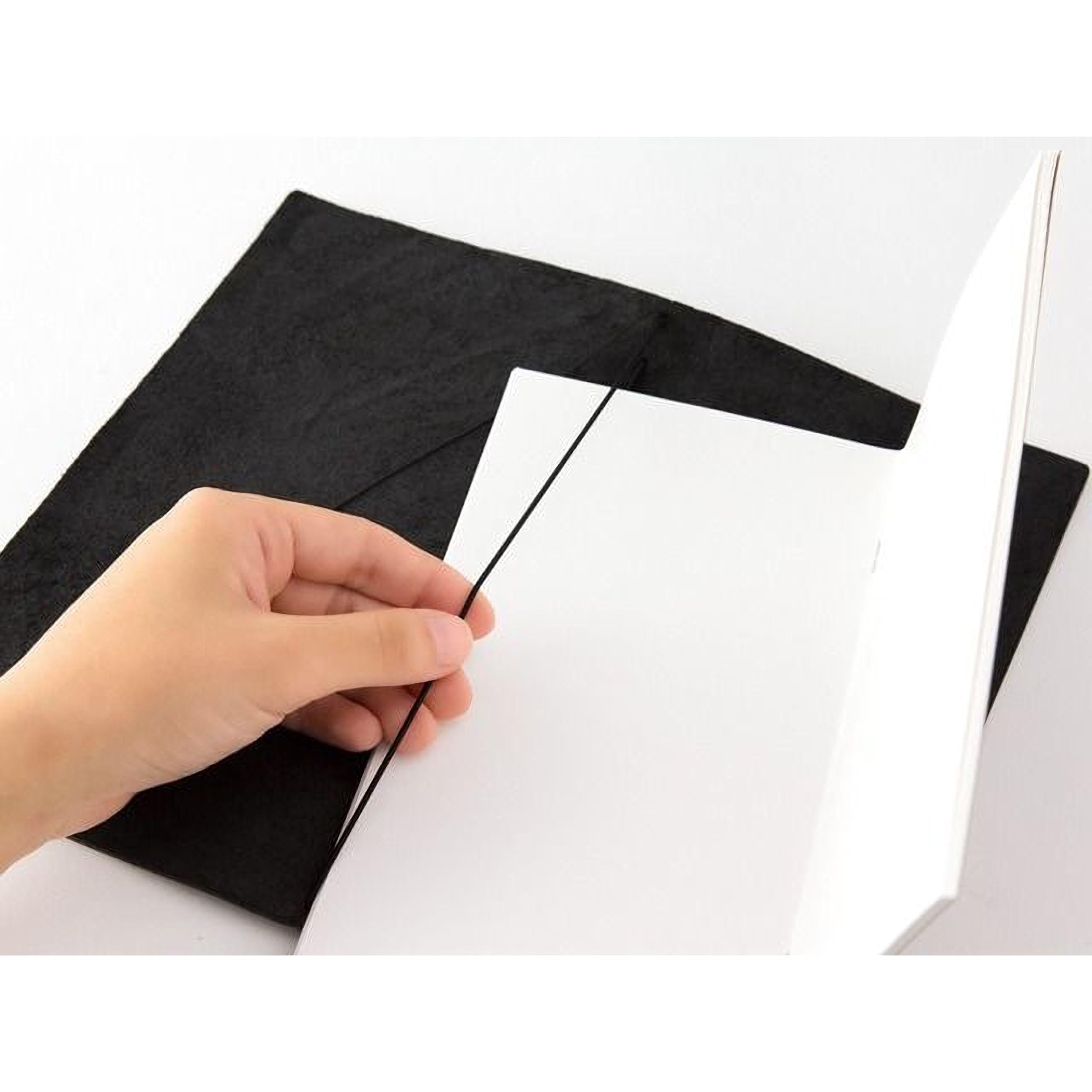 A hand is holding a black object, which appears to be a piece of paper or a notebook, and is positioned over a white surface.