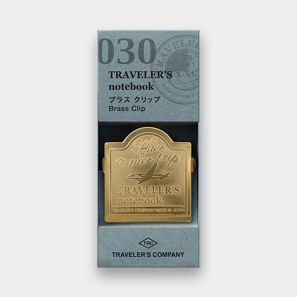 A gold-colored brass clip with the text "Traveler's notebook" printed on it, placed inside a box with a gray and white color scheme.