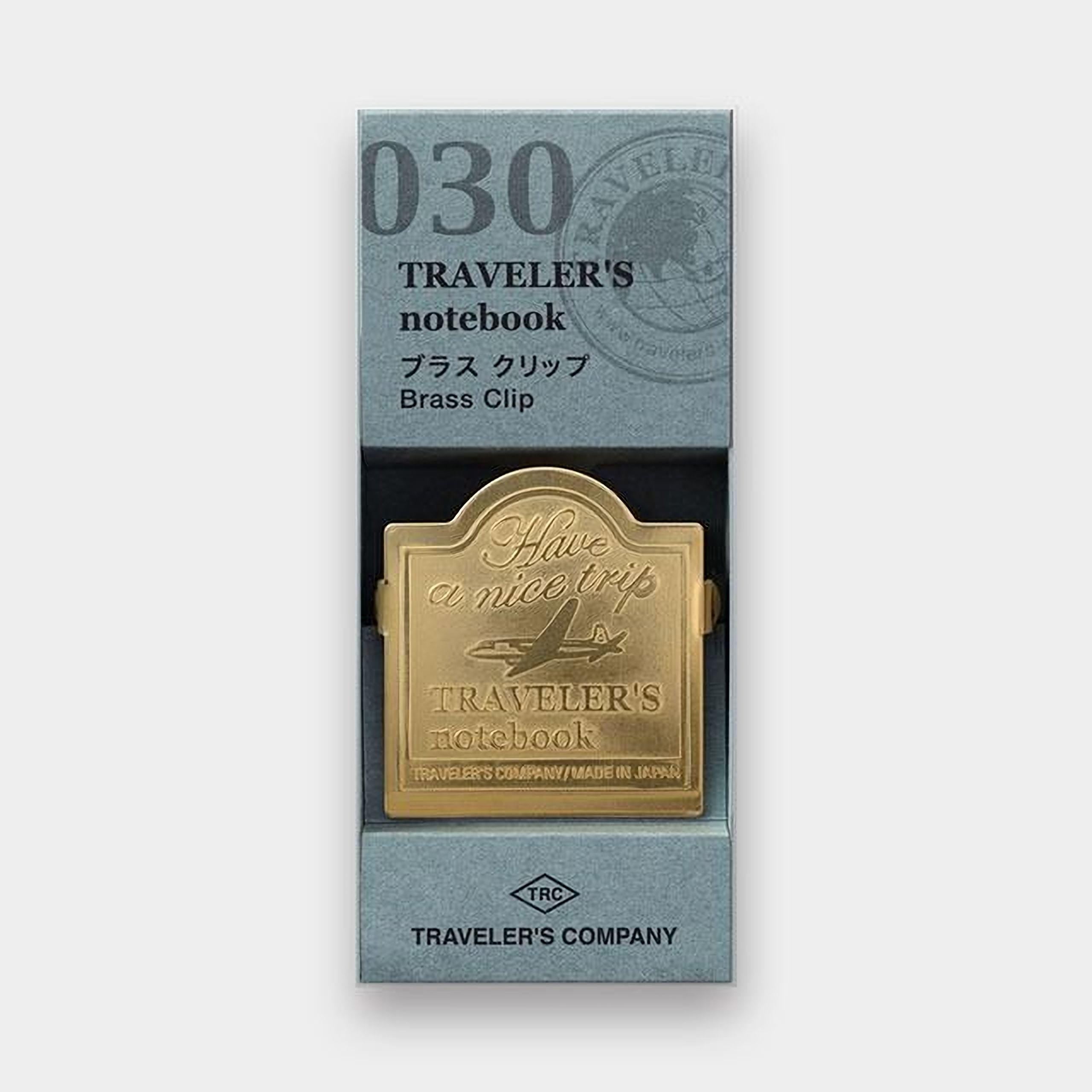 A gold-colored brass clip with the text "Traveler's notebook" printed on it, placed inside a box with a gray and white color scheme.