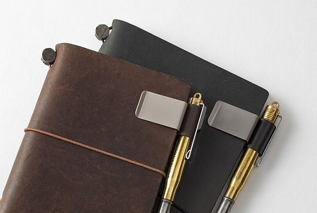 A brown leather-bound notebook, a black notebook, and two pens are arranged on a white surface.