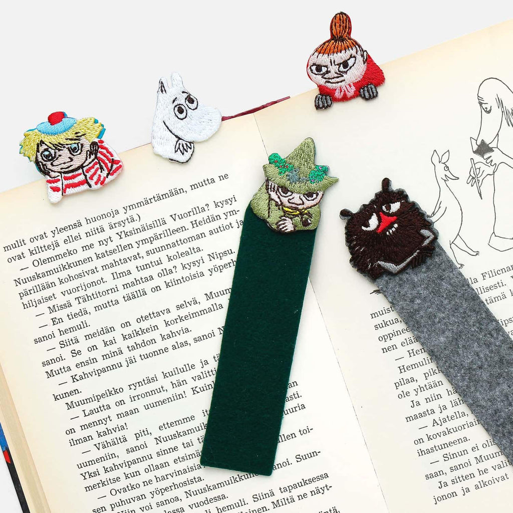 a collection of whimsical, embroidered bookmarks in the shape of characters from the Moomo and the Knights series, including a character with a green hat and a character with a red hat. The bookmarks are placed on top of an open book, adding a playful touch to the reading experience.