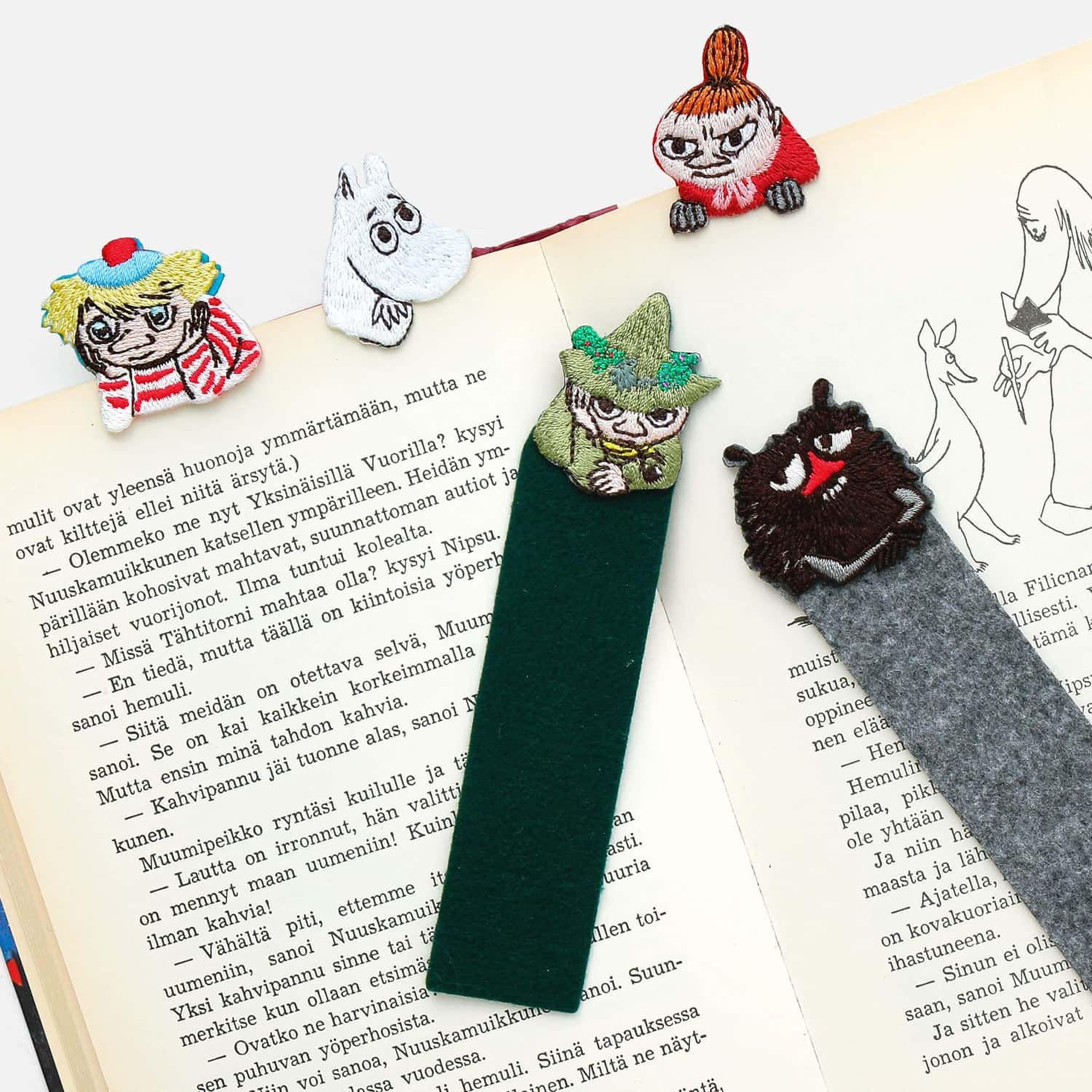a collection of whimsical, embroidered bookmarks in the shape of characters from the Moomo and the Knights series, including a character with a green hat and a character with a red hat. The bookmarks are placed on top of an open book, adding a playful touch to the reading experience.
