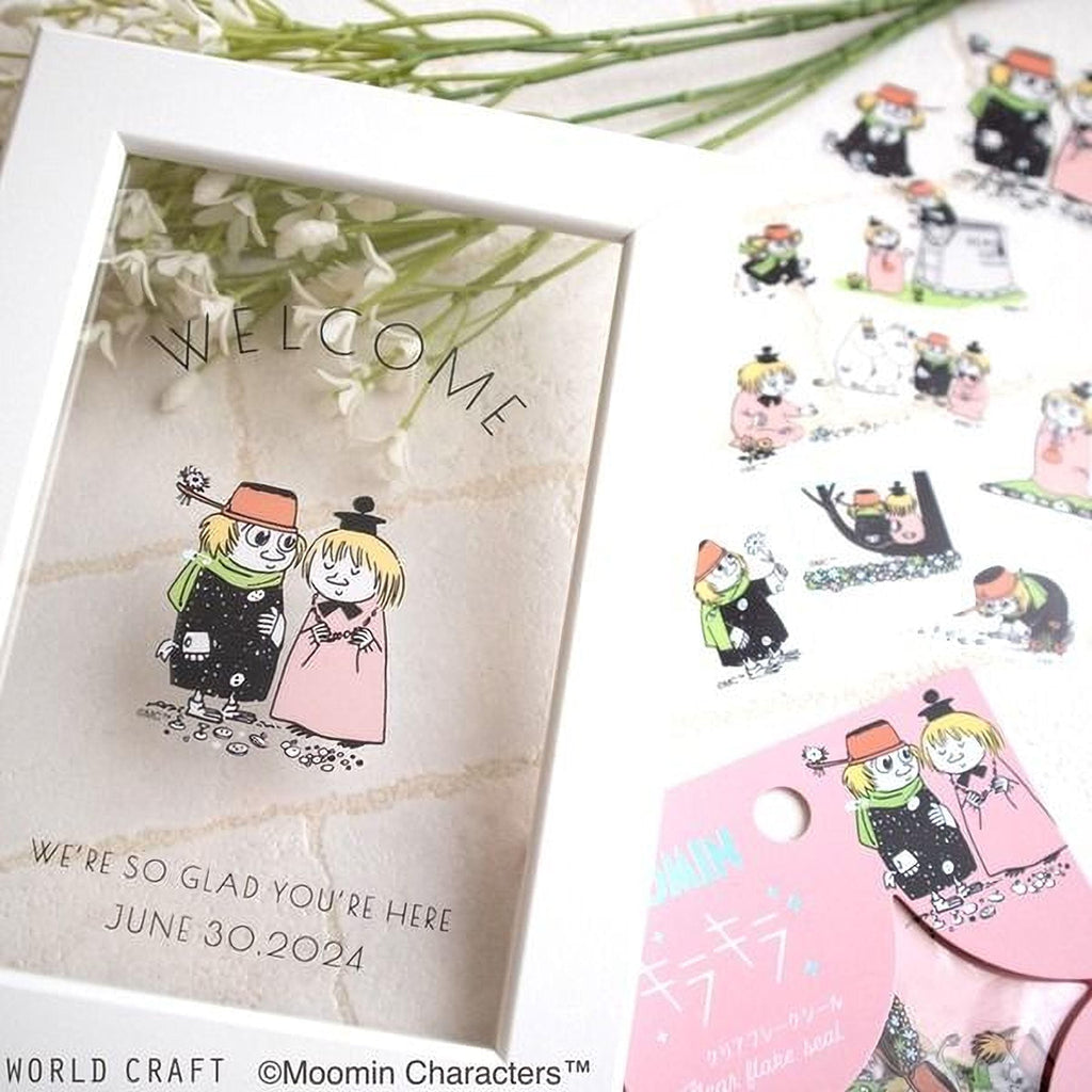 a framed greeting card with illustrations of two people, along with text that reads "Welcome to the world of Moomin characters" and "We're so Glad You're Here June 30, 2024". The card is placed on a table with a bouquet of flowers and other decorative items.