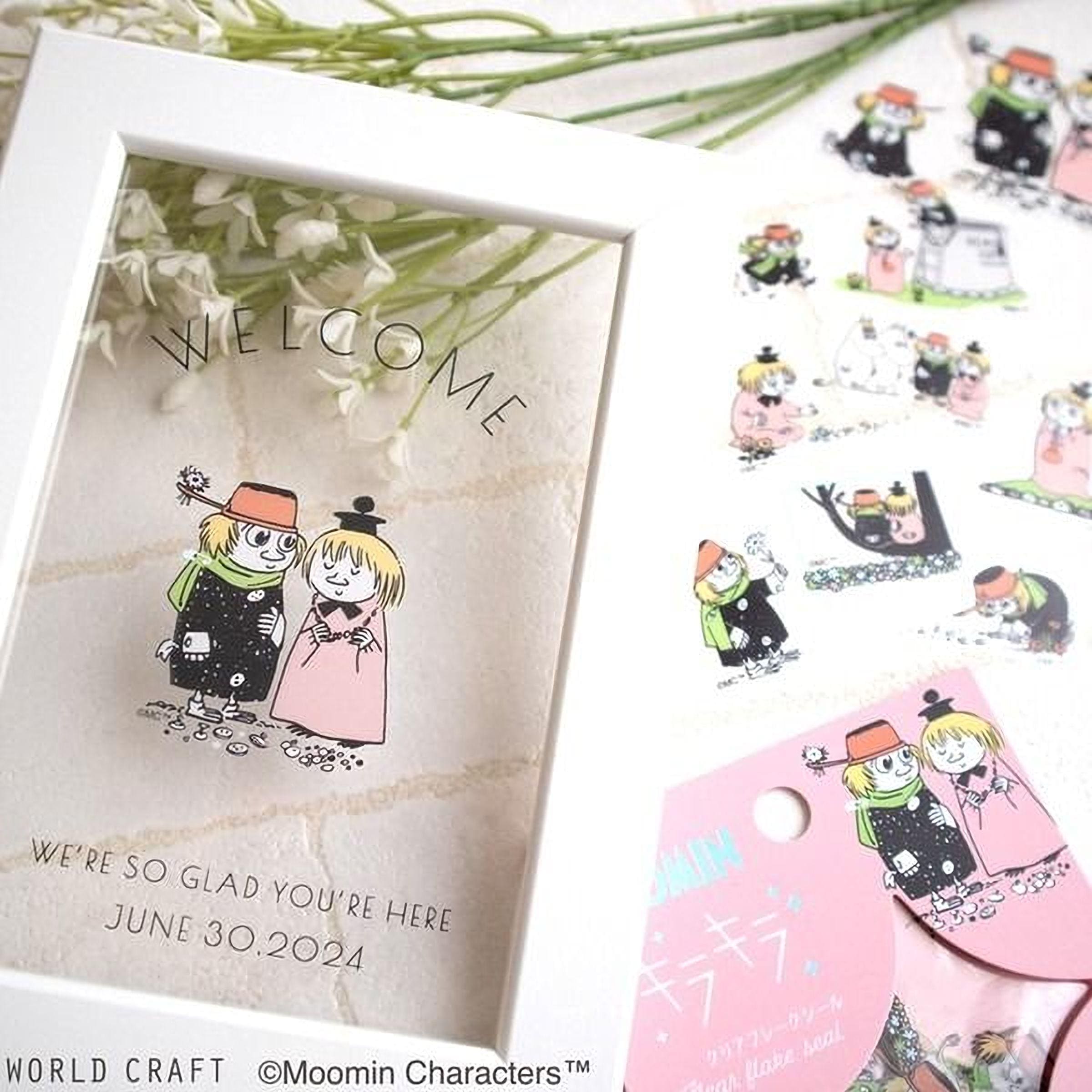 a framed greeting card with illustrations of two people, along with text that reads "Welcome to the world of Moomin characters" and "We're so Glad You're Here June 30, 2024". The card is placed on a table with a bouquet of flowers and other decorative items.